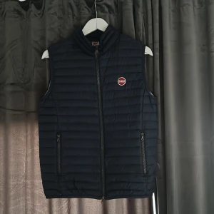 Mörkblå dunväst från Colmar - Snygg mörkblå dunväst från Colmar med dragkedja och broderad logga på bröstet. Västen har en quiltad design och två praktiska fickor med dragkedja framtill. Perfekt nu till våren o sommaren när det blir lite varmare ute. Det är storlek 172 cm ( 16 år ) men sitter som storlek S, säljer i befintlig skick