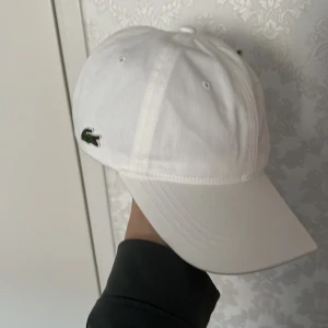 Vit keps från Lacoste - Snygg vit keps från Lacoste med klassisk design och justerbart brunt bälte baktill. Perfekt för en stilren look med den ikoniska krokodilloggan på sidan. Passar till många olika outfits!
