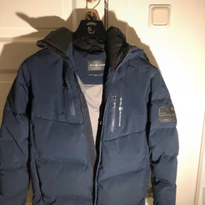 Blå jacka från Sail Racing - Säljer en blå racka från Sail Racing med dragkedja och flera fickor. Perfekt för kalla dagar med sin varma och vadderade design.