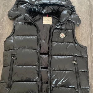 Svart dunväst från Moncler - Snygg svart dunväst från Moncler med glansig finish och dragkedja framtill. Västen har två praktiska fickor med dragkedjor och en klassisk Moncler-logga på bröstet. Perfekt för kyliga dagar när du vill hålla stilen.