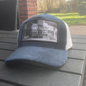 Blå och vit keps med tryck - Snygg keps i blått och vitt med ett coolt tryck av en byggnad på framsidan. Kepsen har ett nätmaterial på baksidan för bättre ventilation och en böjd skärm. Perfekt för en avslappnad stil.