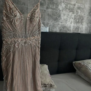 Champagnefärgad klänning från Dressroom Linné - Elegant champagnefärgad långklänning med v-ringad design och detaljerade pärlor och paljetter. Klänningen har en vacker omlott-look och är perfekt för festliga tillfällen. Storlek S/M och använts bara en gång, ordinarie pris 6000.