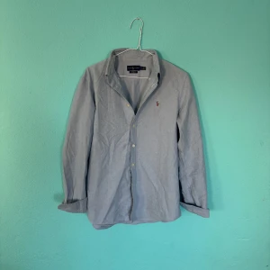 Ljusblå skjorta från Ralph Lauren - Snygg ljusblå skjorta från Ralph Lauren med klassisk krage och broderad logga på bröstet. Skjortan har långa ärmar och knappar framtill. Perfekt för en stilren look.