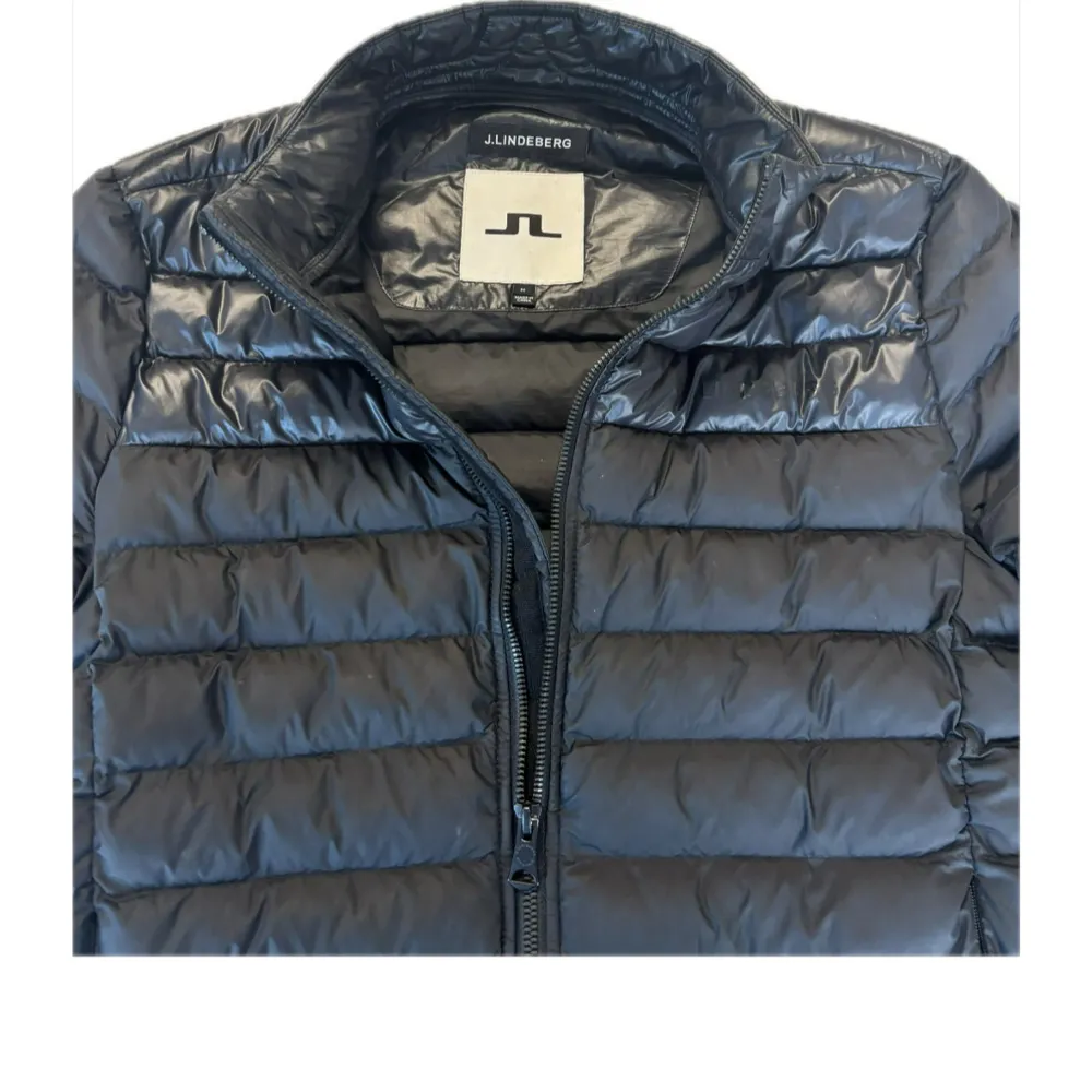 Snygg svart pufferjacka från J.Lindeberg med quiltad design och dragkedja framtill. Jackan har en stilren look med hög glansig krage och långa ärmar. Perfekt till hösten och våren.. Takit.