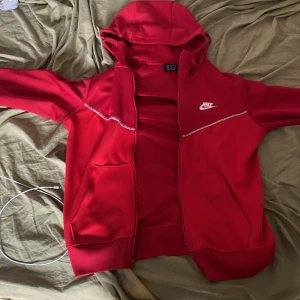 Röd huvtröja från Nike - Säljer en snygg röd huvtröja från Nike med dragkedja och huva. Tröjan har ett vitt Nike-logotyptryck på bröstet och ribbade muddar vid ärmslut och nederkant. Perfekt för en sportig look!