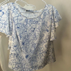 Vit blommig blus från Mohito - Säljer en söt vit blus från Mohito med blått blommönster. Blusen har korta ärmar med volangdetaljer och en rund halsringning. Perfekt för en somrig look! 🌸