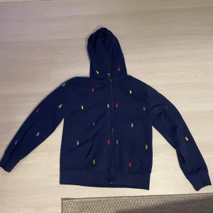 Mörkblå hoodie från Polo Ralph Lauren - Säljer en snygg mörkblå hoodie från Polo Ralph Lauren med färgglada broderade logotyper över hela tröjan. Den har en dragkedja framtill och en praktisk huva. Perfekt för en avslappnad stil. L-14-16= 164