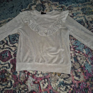 Beige tröja med volangdetalj - Säljer en söt beige tröja med volangdetalj över bröstet. Tröjan har långa ärmar och är perfekt för en stilren look. Materialet ser mjukt och bekvämt ut, vilket gör den idealisk för vardagsbruk.