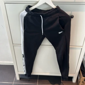Svarta mjukisbyxor från Reebok - Säljer ett par svarta mjukisbyxor från Reebok med vit sidopanel och snörning i midjan. Byxorna har en avslappnad passform och är perfekta för en bekväm stil. Reebok-loggan är broderad på benet.