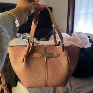 Rosa väska med gulddetalj - Snygg rosa handväska med en elegant gulddetalj framtill. Väskan har två handtag och en justerbar axelrem. Aldrig använd då de va ett inpulsköp.. nypris: 350 kr kom gärna med bud🩷