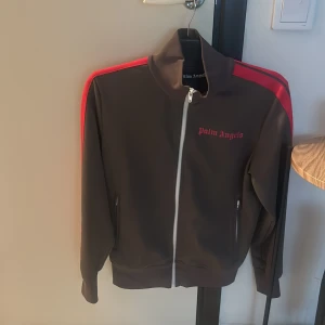 Grå träningsjacka från Palm Angels - Snygg grå träningsjacka från Palm Angels med röda detaljer och dragkedja framtill. Jackan har långa ärmar och en hög krage. Perfekt för en sportig look! Priset kan diskuteras