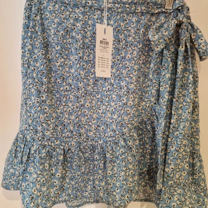 Blommig blå kjol från ONLY stl L - Säljer en söt blå kjol från ONLY med blommigt mönster. Kjolen har volangdetaljer och ett knytband i midjan för en extra touch. Perfekt för en somrig look!