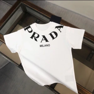 Vit t-shirt från Prada - Snygg vit t-shirt från Prada med stor svart logotyp och texten 'Milano' på framsidan. Perfekt för en stilren och modern look. Kortärmad och tillverkad i mjukt material för extra komfort. Passar strl L för de som pendlar mellan L & XL. annars så är tröjan i strl XL!