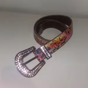 Unikt bälte med färgglatt graffiti-mönster och ett stort, glittrande strasspänne. Perfekt för att ge en extra touch till din outfit.