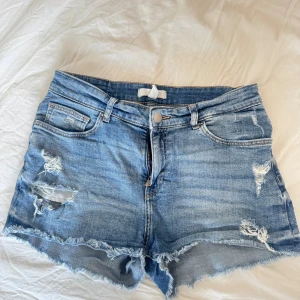 Blå jeansshorts med slitningar - Säljer ett par snygga blå jeansshorts med slitningar och fransiga kanter. De har en klassisk femficksdesign och knappgylf. Perfekta för en avslappnad sommarlook!