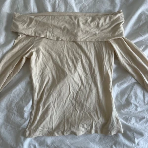 Naturvit offshoulder topp - Snygg beige offshoulder topp med långa ärmar. Perfekt för en stilren look. Materialet är mjukt och följsamt, vilket ger en bekväm passform.