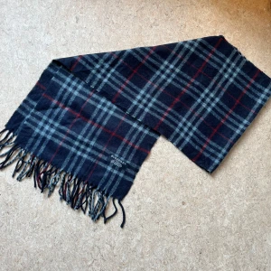 Rutig halsduk från Burberry - Supersnygg och stilren halsduk från Burberry i deras klassiska rutmönster. Den är i mjuk ull och håller dig varm samtidigt som den lyfter vilken outfit som helst. En perfekt accessoar som aldrig går ur stil.  Skick: Väldigt fint begagnat skick – inga synliga defekter. Material: 100 % ull Färg: blå med Burberrys signaturrutor Mått: längd: 140cm           Bred: 30cm  Säljer då den inte kommer till användning längre.