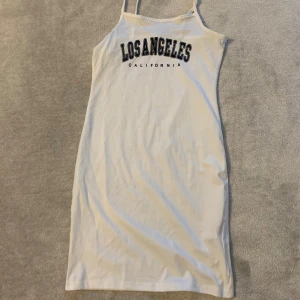 Vit klänning med text - Snygg vit klänning med texten 'Los Angeles California' i svart på framsidan. Klänningen har smala axelband och en enkel design som passar perfekt för varma dagar.