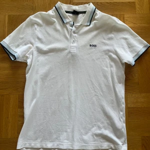 Vit kortärmad pikétröja från Boss - Säljer en stilren vit pikétröja från Boss med korta ärmar och klassisk krage. Tröjan har diskreta svarta och grå detaljer på kragen och ärmsluten samt en broderad logotyp på bröstet. Perfekt för en avslappnad men ändå snygg look. Använd endast ett fåtal gånger. Stl M