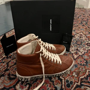  Saint Laurent ”joe sneaker” - Saint Laurent ”joe sneaker”i brunt läder med vit sula och handskriven text: “1971 BAD LIEUTENANT”.. Originalpris låg runt 6 500–7 000 kr när de släpptes, men modellen är inte i produktion. Dem är storlek 41 men passar mer 42!