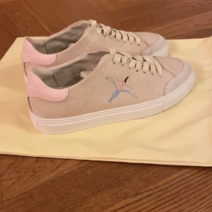 Beige sneakers från Axel Arigato - Snygga beige sneakers från Axel Arigato med rosa detaljer och broderad fågel på sidan. Endast använda ett fåtal gånger. 2600 nypris