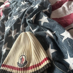 Vit ribbad mössa från Moncler - Snygg vit ribbad mössa från Moncler med broderad logga framtill. Mössan har röd och svart randdetalj vid kanten, perfekt för att hålla värmen med stil.