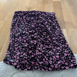 Svart blommig kjol från Divided, Hm  - Säljer en svart kjol med rosa blommigt mönster från Divided. Kjolen har en elastisk midja och är perfekt för en färgglad look. Den är gjord i ett mjukt och bekvämt material. Har rynk mitt fram och bak. 