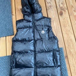 Svart dunväst från Moncler - Snygg svart dunväst från Moncler med glansig yta och dragkedja framtill. Västen har en hög krage och två fickor med dragkedjor. Perfekt för kyliga dagar.