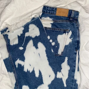Blå jeans med vita fläckar - St W32 - Säljer ett par unika blå jeans från Monki med ett coolt blekt mönster i vitt. De har en hög midja och klassisk femficksdesign. Perfekta för att ge din outfit en edgy touch!