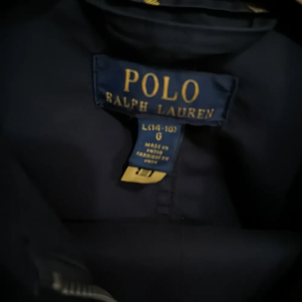 Säljer en stilren marinblå jacka från Polo Ralph Lauren. Jackan har en dragkedja framtill och en klassisk krage. Den är långärmad och har en röd logotyp broderad på bröstet. Perfekt för en snygg och avslappnad stil. Tunn jacka med storleken 14/16 år. Takit.