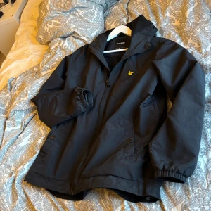 Svart jacka från Lyle & Scott - Snygg svart jacka från Lyle & Scott med dragkedja och huva. Perfekt för kyligare dagar. Jackan har långa ärmar och elastiska muddar för extra komfort. Den klassiska loggan syns på bröstet.