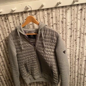 Grå cardigan - Snygg grå cardigan från John ricci. Jackan har en dragkedja framtill och en praktisk huva med snörning. Perfekt för kyliga dagar!