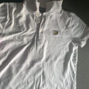 Vit pikétröja från Lyle & Scott - Säljer en stilren vit pikétröja från Lyle & Scott med klassisk krage och knappar framtill. Tröjan har en broderad logotyp på bröstet och är kortärmad. Perfekt för en avslappnad och snygg look.