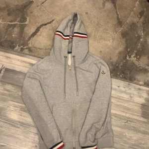 Grå Moncler hoodie - Säljer en stilren grå hoodie från Moncler med dragkedja och justerbar huva. Den har Monclers logga på ärmen och ribbade muddar. Perfekt för en avslappnad stil. Säljer denna äkta moncler kofta för 2000kr då den har några skador vid armen. Nypris är ca 5600kr. Pris kan diskuteras. Tar även emot byten.