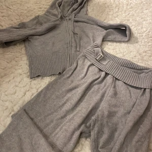 Grå croppad hoodie med matchande byxor - Säljer en grå croppad hoodie med dragkedja och matchande byxor. Hoodien har långa ärmar och ribbade detaljer. Byxorna har en bred ribbad midja med bälte. Perfekt för en avslappnad stil.