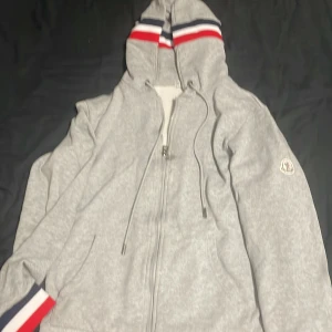 Grå hoodie från Moncler - Säljer en stilren grå hoodie från Moncler med dragkedja och broderad logga på ärmen. Huvan har ränder i rött, vitt och blått som ger en sportig touch. Perfekt för kyliga dagar!