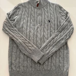 Grå stickad tröja från Polo Ralph Lauren - Säljer en stilren grå stickad tröja från Polo Ralph Lauren med kabelstickat mönster och dragkedja vid halsen. Tröjan har långa ärmar och en broderad logotyp på bröstet. Perfekt för kyligare dagar!