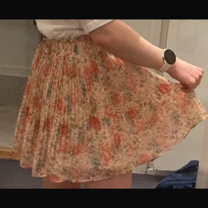 Blommig kortkjol med volang - Charmig blommig kortkjol i ljusa färger med volangdetaljer. Perfekt för sommarens varma dagar. Kjolen har ett vackert blommönster i orange och grönt på en ljus bas.