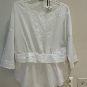 Vit broderad blus med trekvartsärm - Superfin vit blus med broderade detaljer framtill och längs midjan. Blusen har trekvartsärmar och en lös passform med lätt transparent tyg. Perfekt för dig som gillar romantiska och luftiga plagg med snygga detaljer.