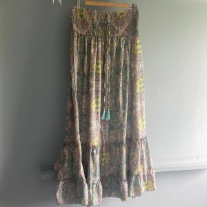 Färgglad långkjol från Bigg Bull - Supersöt långkjol från Bigg Bull med färgglatt blommigt mönster i pastelliga toner av rosa, blått, gult och grönt. Kjolen har smock i midjan, knytband med tofsar och volang nertill. sälj dp den inte används längre