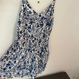 Blommig playsuit från H&M devided - Playsuit med blått blommönster på vit botten. Playsuiten har smala axelband, v-ringning och markerad midja. Säljer då den är liten.
