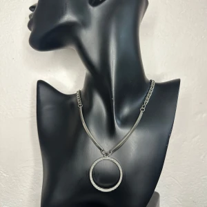 Silverfärgat halsband med cirkelhänge -  Vintagehalsband från 1970-talet i kraftfull brutaliststil med böjda metallstavar och stort cirkelformat hänge. Ca 47 cm långt, hänget är ca 4 cm. Ytan är silvrig, och det finns otydliga stämplar som eventuellt tyder på silver – men osäkert så säljs därför inte som silver. Ett uttrycksfullt smycke som passar både minimalistisk och alternativ stil. Prutning undanbedes, men jag erbjuder bundlerabatt.  Jag rensar smyckesskrin och garderob – kolla gärna mina andra annonser också!