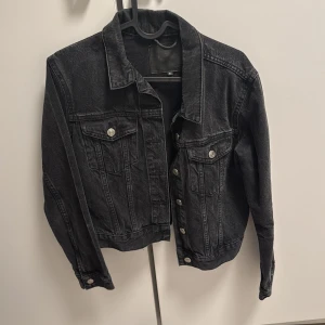 Svart jeansjacka från 157, stl S - Svart jeansjacka från 157 i klassisk modell med silvriga knappar framtill och på bröstfickorna. Jackan har en rak passform, krage och långa ärmar. Perfekt att slänga över en hoodie eller t-shirt för en chill look.