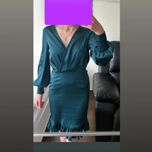 Mörkgrön omlottklänning med volang - Supersnygg mörkgrön omlottklänning i satinliknande material med v-ringning, långa puffärmar och smock vid ärmslut. 