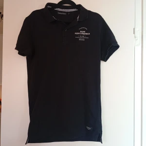 Svart pikétröja Peak Performance S - Svart pikétröja från Peak Performance i storlek S. Klassisk krage, korta ärmar och vit logga tryckt på bröstet. Snygg detalj med siffran 86 på ena ärmen. Tröjan har en stilren, sportig look.