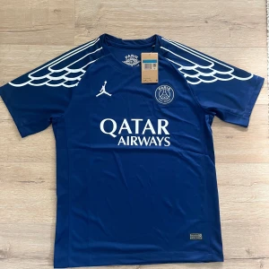 PSG x Jordan blå fotbollströja - Blå PSG fotbollströja från Jordan med vita detaljer och vingmönster på axlarna. Tröjan har korta ärmar, rund hals och är tillverkad i lätt, ventilerande material. PSG-logga och Jumpman-logga på bröstet samt Qatar Airways-tryck framtill.