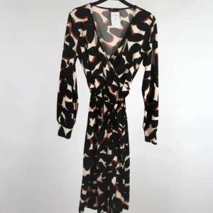 Omlottklänning med grafiskt mönster från Stockh LM, Nyskick, stl XS/34 - Elegant omlottklänning från Stockh LM i svart, benvitt och rostbrunt grafiskt mönster. Smickrande passform med knytning i midjan, puffärm och bred manschett. Mjuk och stretchig kvalitet (95 % polyester, 5 % elastan). Storlek XS, men passar även S tack vare omlottdesignen. I mycket fint skick – ser oanvänd ut. Ingen prutning – men jag erbjuder bundlerabatt. Rensar garderoben, så kika gärna på mina andra annonser också!  ⸻  