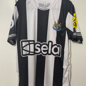 Newcastle United Isak 14 matchtröja - Säljer en Newcastle United fotbollströja med Isak 14 på ryggen. Tröjan är svartvit med breda ränder, korta ärmar och flera sponsorloggor, bland annat Premier League och Noon. Materialet är lätt och andas, perfekt för match eller träning. Plus att tröjan är helt ny och aldrig använt.