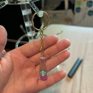 Lila Starbucks flask-nyckelring - Nyckelring med en liten flaska i lila och beige nyans, dekorerad med Starbucks-logga. Flaskan hänger i en guldfärgad kedja och ring. Perfekt accessoar för dig som älskar kaffe och vill ha något unikt på väskan eller nycklarna.