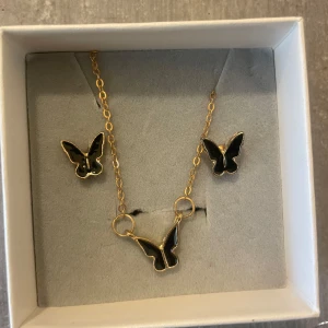 Guldigt fjärilssmyckeset svart - Set med halsband och örhängen i guldtonad metall med svarta fjärilar. Halsbandet har en tunn kedja och en fjäril i mitten, matchande örhängen med samma fjärilsmotiv. Perfekt för dig som gillar trendiga och söta detaljer.